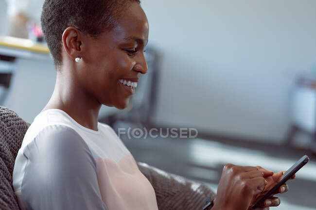 Femme d'affaires afro-américaine souriante assise dans un fauteuil, à l'aide d'un smartphone. entreprise créative indépendante dans un bureau moderne. — Photo de stock