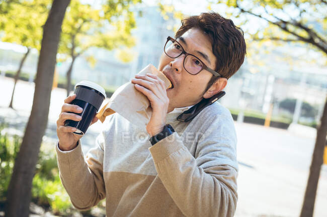 Asian businessman using eating sandwich and holding takeaway coffee sitting in city street. businessman on the go out and about in city concept. — Stock Photo