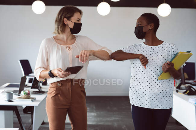 Zwei unterschiedliche Geschäftsfrauen tragen Gesichtsmasken, halten Tabletten und Dokumente in der Hand, berühren Ellbogen. Selbstständiges kreatives Geschäft in einem modernen Büro während der Coronavirus-Pandemie 19. — Stockfoto