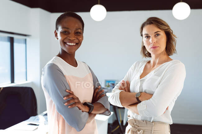 Due diverse donne d'affari sorridenti in piedi con le braccia incrociate al lavoro. affari creativi in un ufficio moderno. — Foto stock