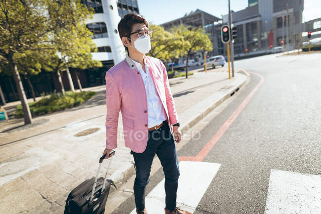 Asian businessman wearing face mask walking with suitcase crossing city street. business travel out and about in city during covid 19 pandemic concept. — Stock Photo