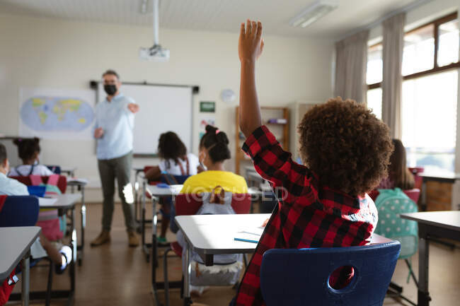 Вид сзади на африканского американского мальчика, поднимающего руку, сидящего на столе в начальной школе. гигиена и социальное дистанцирование в школе во время пандемии ковида 19 — стоковое фото