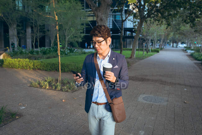 Asiatischer Geschäftsmann mit Smartphone hält Kaffee zum Mitnehmen in der Straße. Digitaler Nomade im Stadtkonzept unterwegs. — Stockfoto