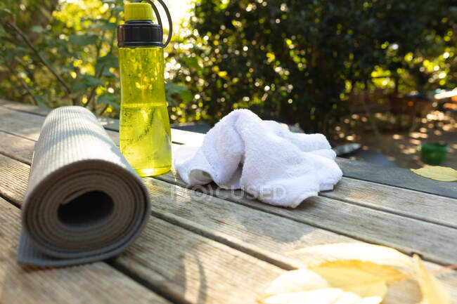 Vista del tappeto yoga arrotolato, asciugamano bianco e bottiglia d'acqua gialla sulla terrazza. accessori fitness e stile di vita attivo. — Foto stock