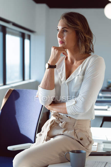 Retrato de una mujer de negocios caucásica pensativa de pie, mirando por la ventana. negocio creativo independiente en una oficina moderna. - foto de stock