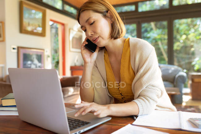 Mulher asiática falando no smartphone e usando laptop, trabalhando em casa. em casa em isolamento durante o confinamento de quarentena. — Fotografia de Stock