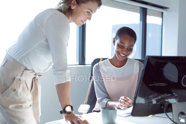Dos empresarias sonrientes diversas que trabajan juntas usando el ordenador portátil, hablando. negocio creativo independiente en una oficina moderna. - foto de stock