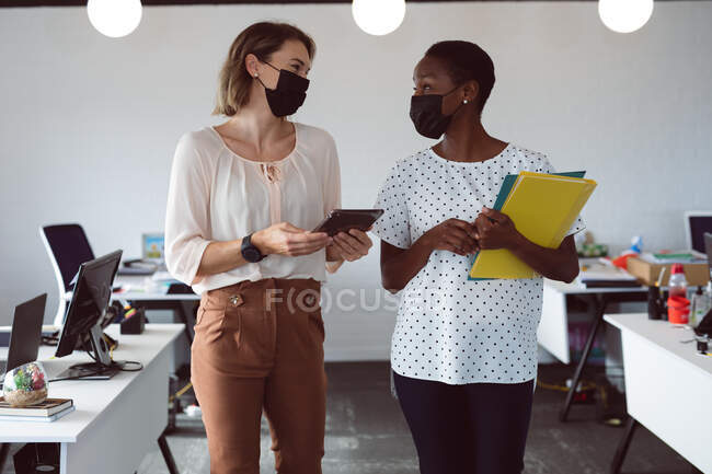 Дві різні бізнес-леді носять маски для обличчя, тримають планшет і документи, розмовляють. незалежний творчий бізнес в сучасному офісі під час пандемії коронавірусу 19 . — стокове фото