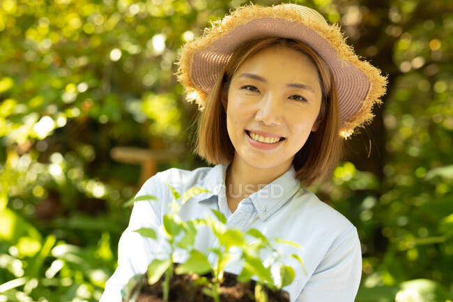 Sorridente donna asiatica indossando cappello di paglia e tenendo pianta in giardino. a casa isolata durante l'isolamento in quarantena. — Foto stock