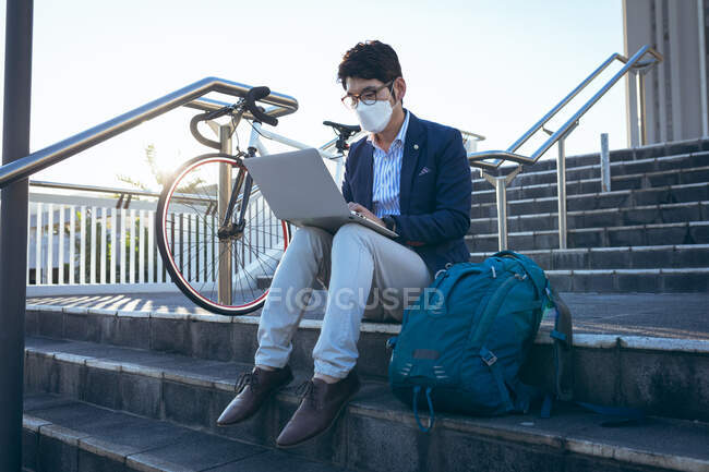 Asian businessman wearing face mask using laptop sitting on steps in city street. digital nomad out and about in city during covid 19 pandemic concept. — Stock Photo