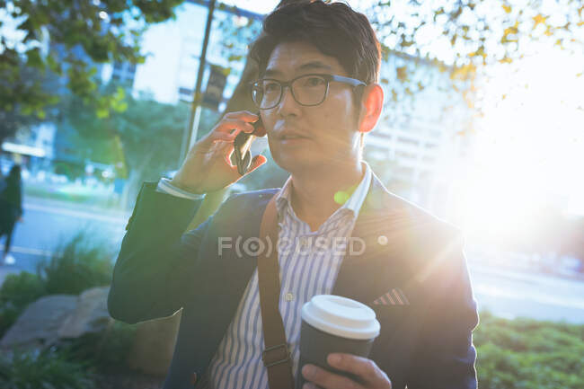 Asiatischer Geschäftsmann, der auf einem Smartphone Kaffee zum Mitnehmen hält, läuft durch die Straßen der Stadt. Digitaler Nomade im Stadtkonzept unterwegs. — Stockfoto