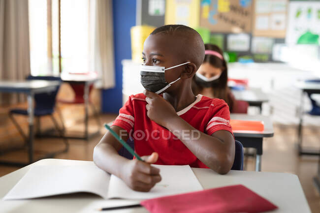 Ein afroamerikanischer Junge mit Mundschutz sitzt in der Klasse der Grundschule auf seinem Schreibtisch. Hygiene und soziale Distanzierung in der Schule während der covid 19 Pandemie — Stockfoto
