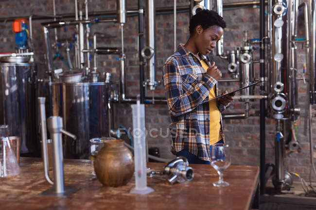 Thoughtful african american female worker holding pen and clipboard at gin distillery. alcohol production and filtration concept — Stock Photo