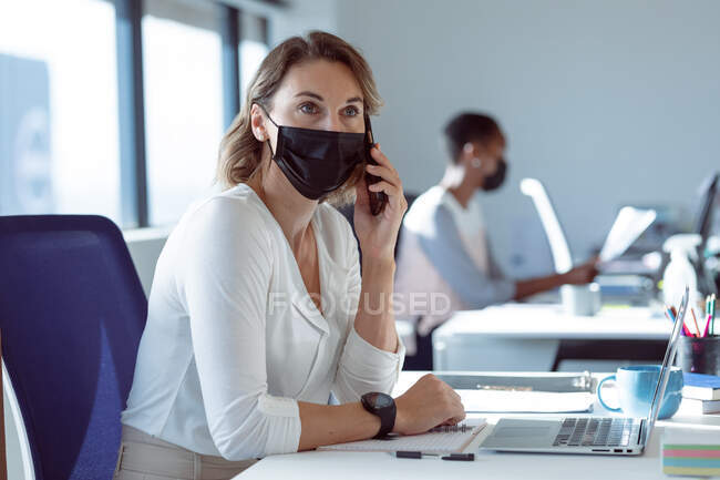 Mulher de negócios caucasiana usando máscara facial, sentada na mesa, usando smartphone. negócio criativo independente em um escritório moderno durante coronavírus covid 19 pandemia. — Fotografia de Stock