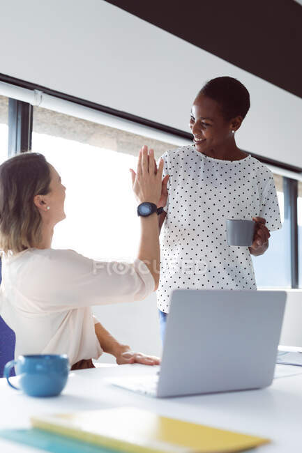 Due donne d'affari diverse alla scrivania, che parlano e danno il cinque. attività creativa indipendente in un ufficio moderno. — Foto stock