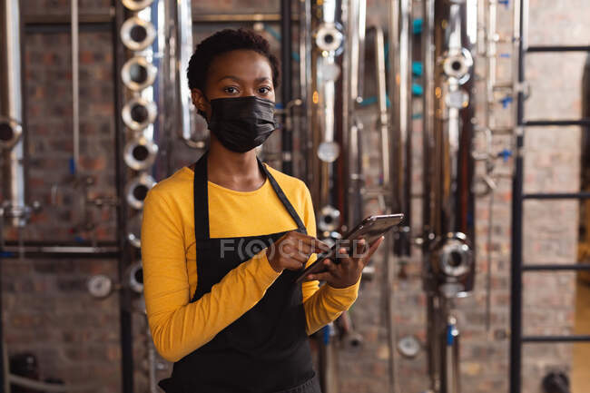 Портрет африканської жінки-робітниці, одягненої в маску обличчя за допомогою цифрового планшета на винокурні джину. Виробництво алкоголю і фільтрація. — стокове фото