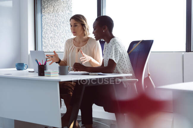 Two diverse businesswomen sitting at desk, looking at laptop, talking. independent creative business at a modern office. — Stock Photo