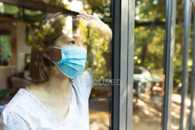 Грустная азиатка в маске и выглядывает в окно. в доме в изоляции во время ковида 19 пандемии и карантинной изоляции. — стоковое фото