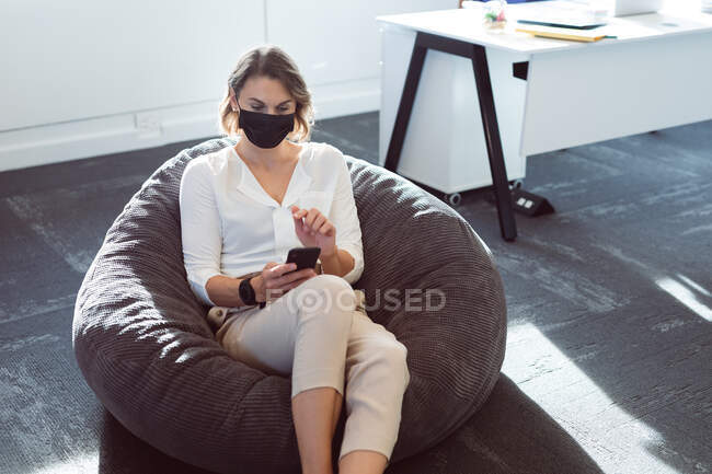 Femme d'affaires caucasienne portant un masque facial, assise dans un fauteuil, utilisant un smartphone. entreprise créative indépendante dans un bureau moderne pendant coronavirus covid 19 pandémie. — Photo de stock