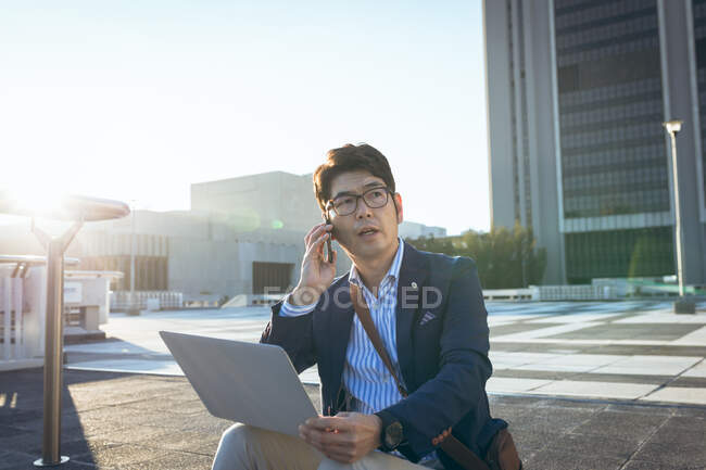 Азиатский бизнесмен в маске с помощью смартфона и планшета сидит на ступеньках на городской улице. Цифровой кочевник в городе во время ковида 19 пандемической концепции. — стоковое фото