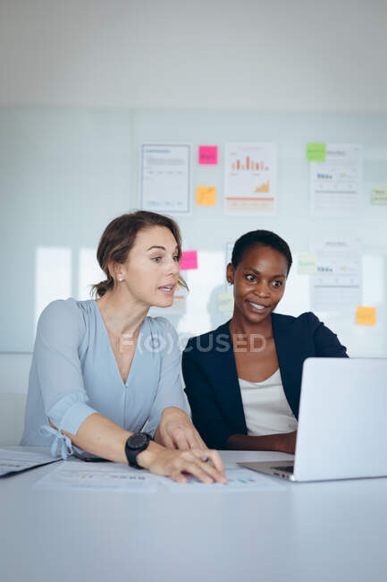 Dos empresarias diversas sentadas en el escritorio, usando el ordenador portátil, hablando. negocio creativo independiente en una oficina moderna. - foto de stock