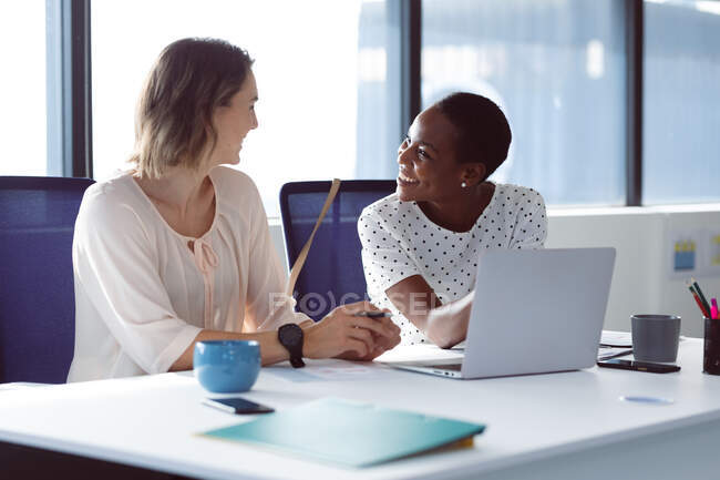 Due diverse donne d'affari sedute alla scrivania, che parlano e sorridono. attività creativa indipendente in un ufficio moderno. — Foto stock