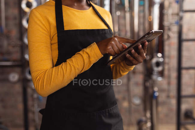 Mid section of african american female worker wearing apron using digital tablet at gin distillery. alcohol production and filtration concept — Stock Photo