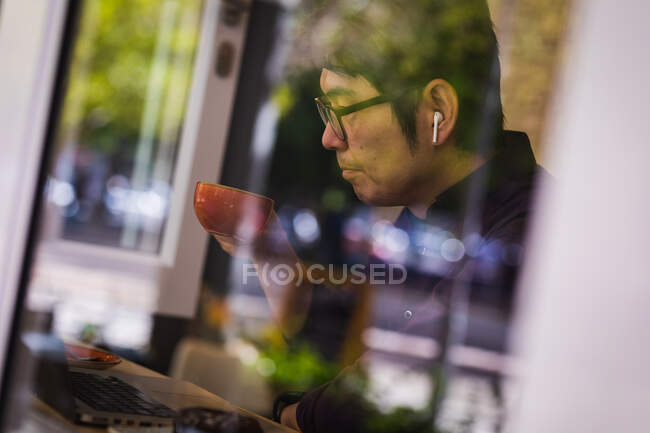 Asian businessman using laptop and wireless earphones drinking coffee in cafe. business travel, digital nomad on the go out and about in city concept. — Stock Photo