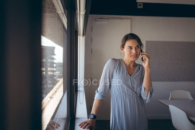 Mulher de negócios caucasiana em pé na janela, falando por smartphone no trabalho. negócio criativo independente em um escritório moderno. — Fotografia de Stock