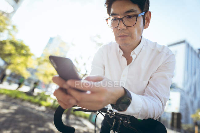 Uomo d'affari asiatico che utilizza smartphone appoggiato sulla bicicletta in strada. nomade digitale in giro per la città concetto. — Foto stock