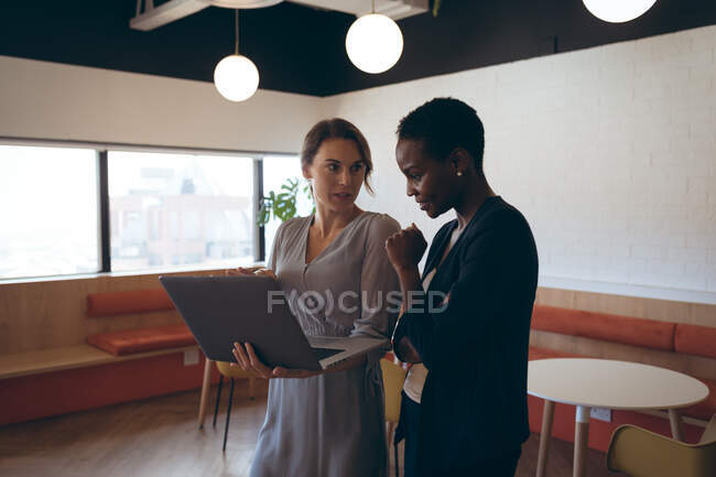 Дві різні бізнес-жінки стоять, розмовляють, використовуючи ноутбук на роботі. Незалежний творчий бізнес у сучасному офісі.. — стокове фото