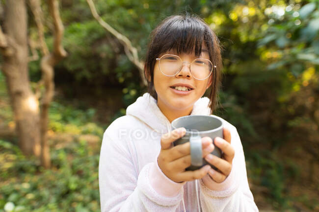 Portrait de fille asiatique souriante en sweat à capuche rose tenant une tasse de thé debout dans le jardin. à domicile en isolement pendant le confinement en quarantaine. — Photo de stock
