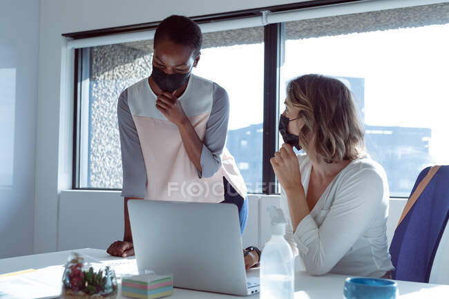Two diverse smiling businesswomen wearing face masks, working together, using laptop, talking. independent creative business at a modern office during coronavirus covid 19 pandemic. — Stock Photo