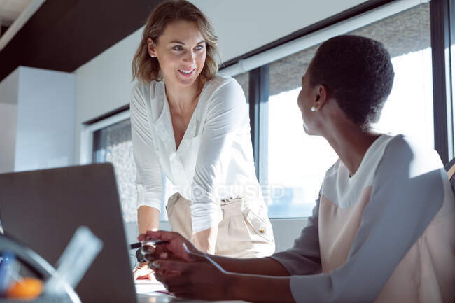 Две разноплановые улыбающиеся деловые женщины работают вместе, используют ноутбук, разговаривают. независимый творческий бизнес в современном офисе. — стоковое фото