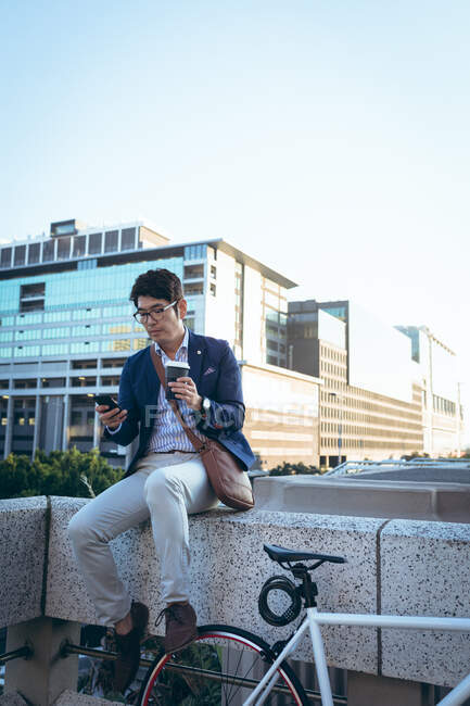 Hombre de negocios asiático usando un teléfono inteligente sosteniendo café para llevar sentado junto a su bicicleta en la calle de la ciudad. nómada digital en concepto de ciudad. - foto de stock
