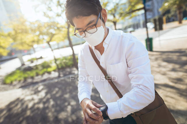 Asian businessman wearing face mask using smartwatch in city street. digital nomad out and about in city during covid 19 pandemic concept. — Stock Photo