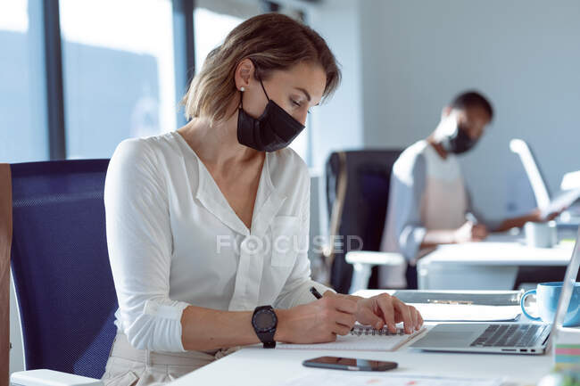 Empresaria caucásica con máscara facial, sentada en el escritorio, tomando notas. negocio creativo independiente en una oficina moderna durante coronavirus covid 19 pandemia. - foto de stock