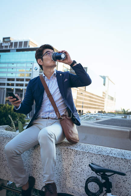 Азиатский бизнесмен, использующий смартфон, пьющий кофе на вынос, сидит на велосипеде на городской улице. цифровая реклама и концепция города. — стоковое фото