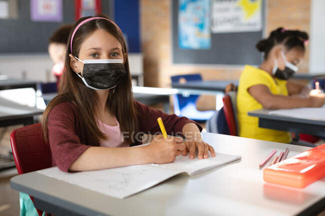 Ritratto di ragazza caucasica che indossa una maschera per il viso seduta sulla scrivania in classe alle elementari. igiene e distanza sociale a scuola durante la covd 19 pandemia — Foto stock