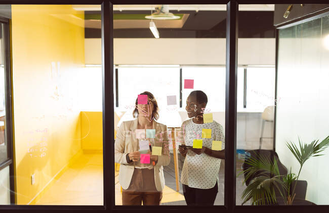 Two diverse businesswomen standing at transparent board, making notes and talking. independent creative business at a modern office. — Stock Photo