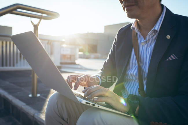 Мидсекция азиатского бизнесмена с помощью ноутбука сидит на ступеньках на городской улице. цифровая реклама и концепция города. — стоковое фото