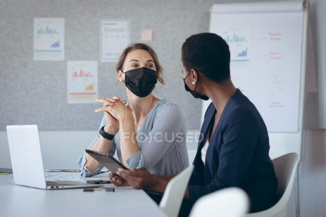 Две разные деловые женщины в масках, сидят за столом, используют ноутбук, разговаривают. независимый творческий бизнес в современном офисе во время пандемии коронавируса. — стоковое фото