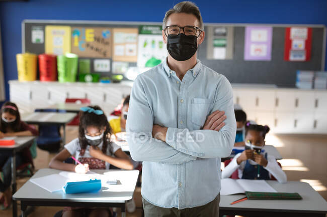 Porträt eines kaukasischen männlichen Lehrers mit Mundschutz, der in der Klasse der Grundschule steht. Hygiene und soziale Distanzierung in der Schule während der covid 19 Pandemie — Stockfoto