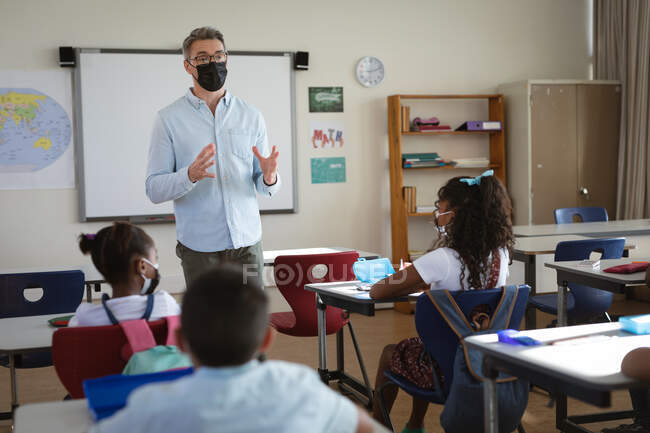 Maestra caucásica con máscara facial enseñando en la clase en la escuela primaria. higiene y distanciamiento social en la escuela durante la pandemia de covid 19 - foto de stock