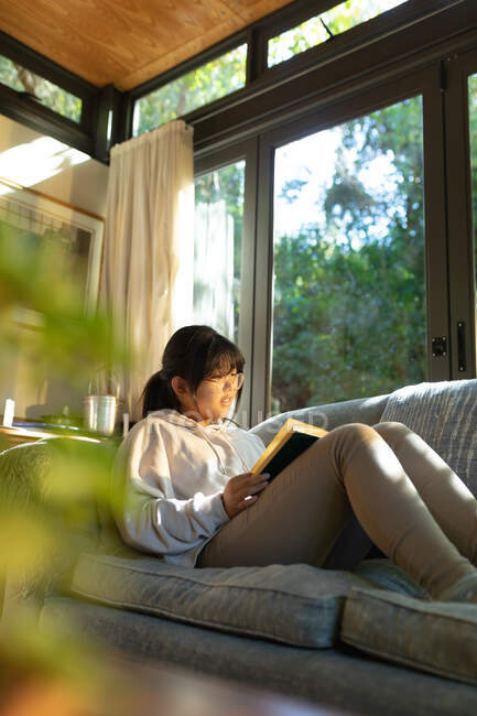 Улыбающаяся азиатка в очках читает книгу и сидит на диване. в доме в изоляции во время карантинной изоляции. — стоковое фото