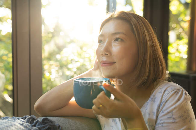 Retrato de mujer asiática sonriente sosteniendo taza de té y sentado en el sofá. en casa en aislamiento durante el bloqueo de cuarentena. - foto de stock