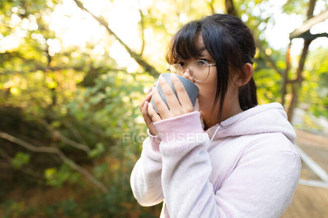 Asiatisches Mädchen in rosa Kapuzenpulli, das Tee trinkt, steht im Garten. Zuhause in Isolation während der Quarantäne. — Stockfoto