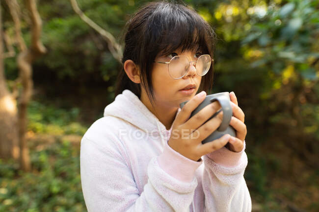 Portrait de fille asiatique en sweat à capuche rose tenant une tasse de thé debout dans le jardin. à domicile en isolement pendant le confinement en quarantaine. — Photo de stock