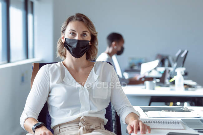 Mujer de negocios caucásica con máscara facial, sentada en el escritorio, mirando a la cámara. negocio creativo independiente en una oficina moderna durante coronavirus covid 19 pandemia. - foto de stock