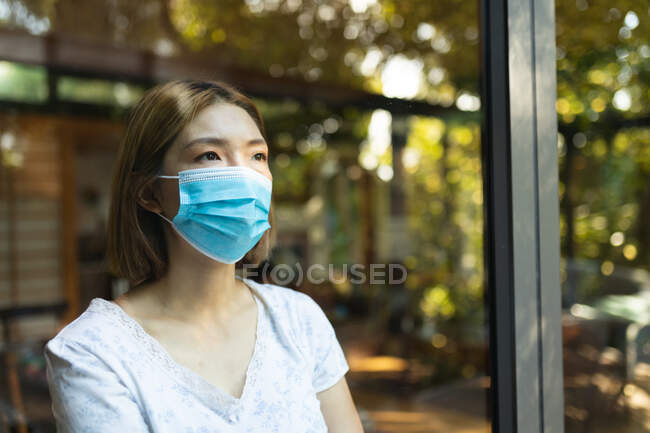 Sad asian woman wearing face mask and looking out of window. at home in isolation during covid 19 pandemic and quarantine lockdown. — Stock Photo